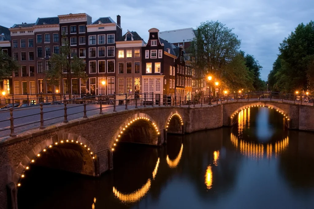 Il canale Reguliersgracht all'incrocio con il Keizersgracht al crepuscolo: ponti ad arco illuminati che si riflettono nell'acqua e, sullo sfondo, case strette affacciate sul canale