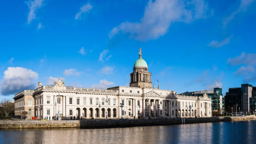 La Custom House di Dublino, edificio neoclassico con cupola verde, vista dall'altra sponda del fiume Liffey sotto un cielo azzurro