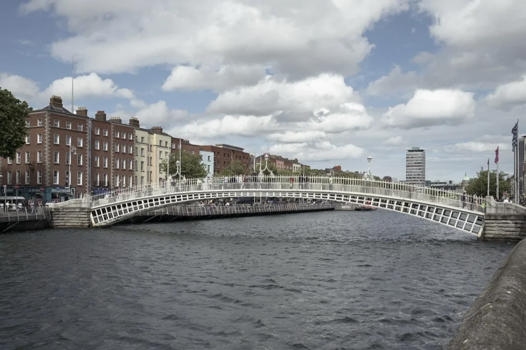 L'Ha'penny Bridge, ponte pedonale bianco in ghisa sul fiume Liffey a Dublino, con edifici in mattoni sulla riva e cielo nuvoloso