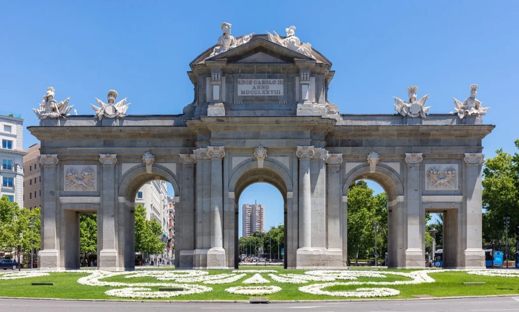 La Puerta de Alcalá con le aiuole fiorite davanti, sotto un cielo azzurro