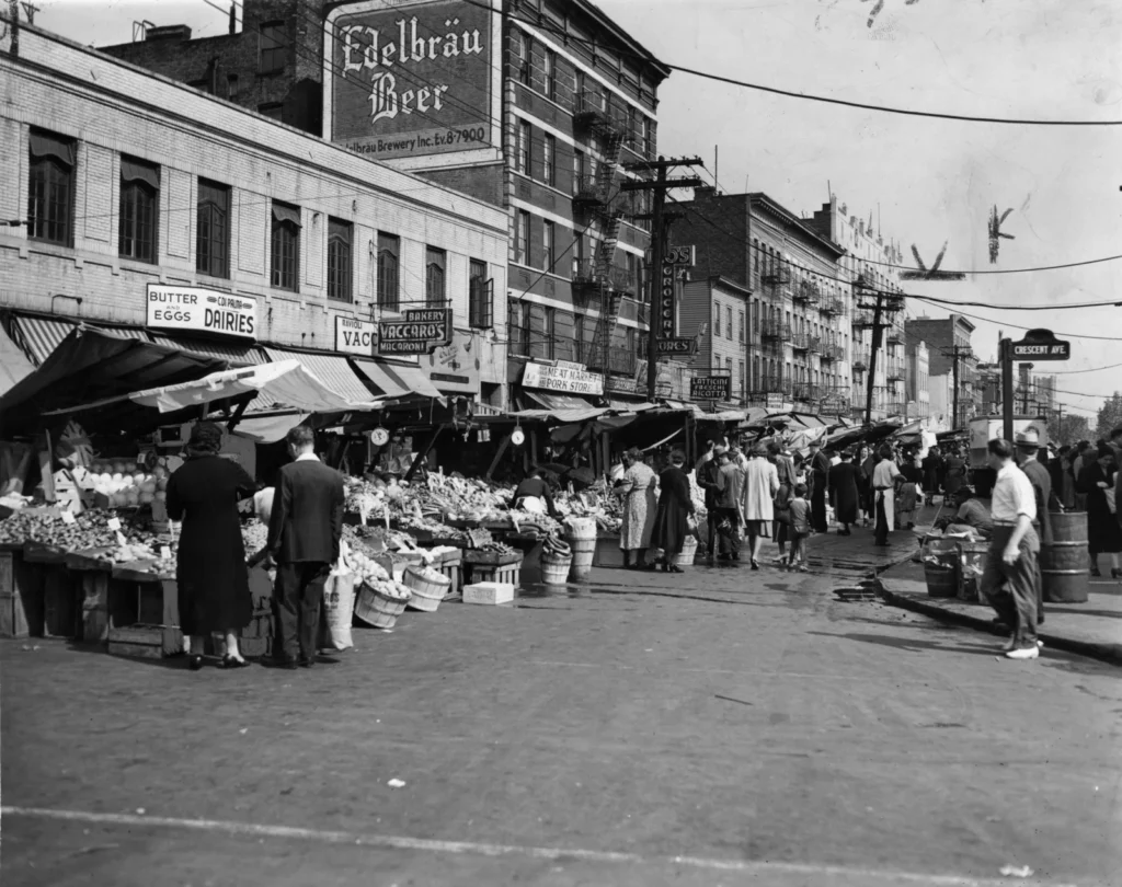 Foto in bianco e nero del 1940: banchi di frutta e verdura su Arthur Avenue, nel Bronx, con le insegne Latticini Freschi e Vaccaro's Bakery e passanti con cappotti e cappelli