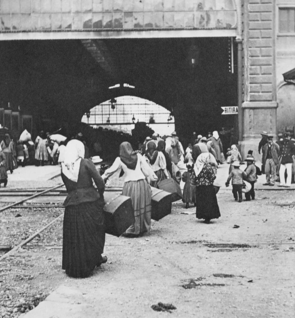 Donne con lo scialle in testa, di spalle, portano bauli verso la tettoia dei binari della stazione; intorno bambini e uomini con il cappello
