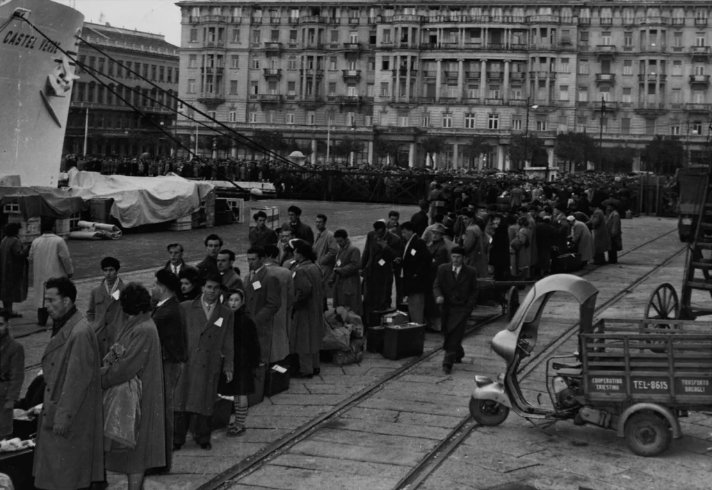 Emigranti in cappotto, con valigie e cartellini appuntati sui vestiti, in fila sulla banchina del porto di Trieste accanto alla prua della nave Castel Verde
